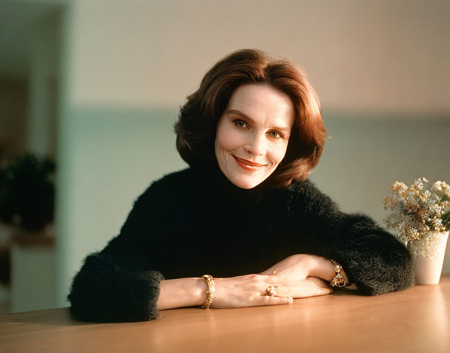 Smiling woman with brown hair in black turtleneck and gold jewelry by table with white bouquet