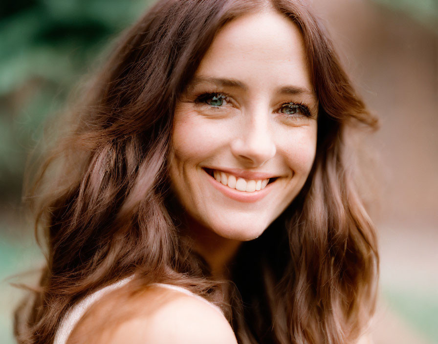 Smiling woman with long wavy brown hair and hazel eyes outdoors in soft-focus green background