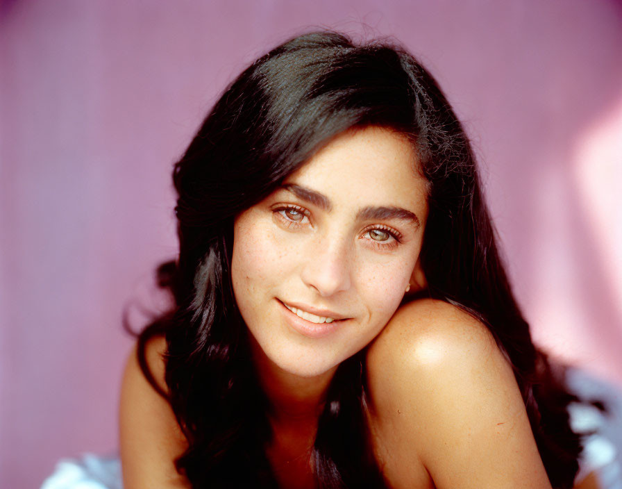 Smiling young woman with dark hair and brown eyes in light blue top on pink background