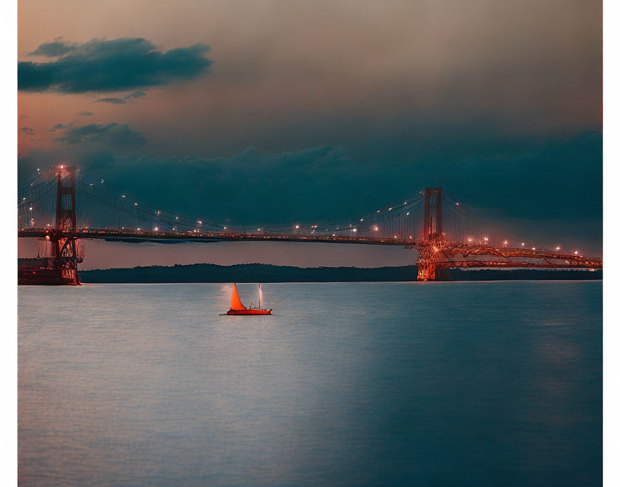 Orange sail sailboat on calm waters at dusk with illuminated suspension bridge.