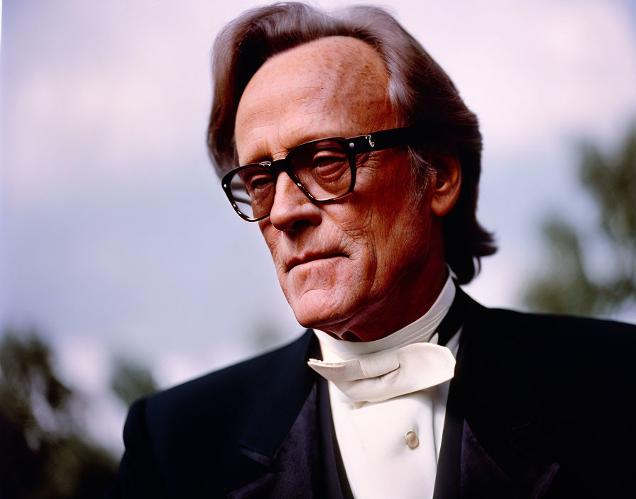 Elderly man in formal tuxedo and glasses gazes thoughtfully.