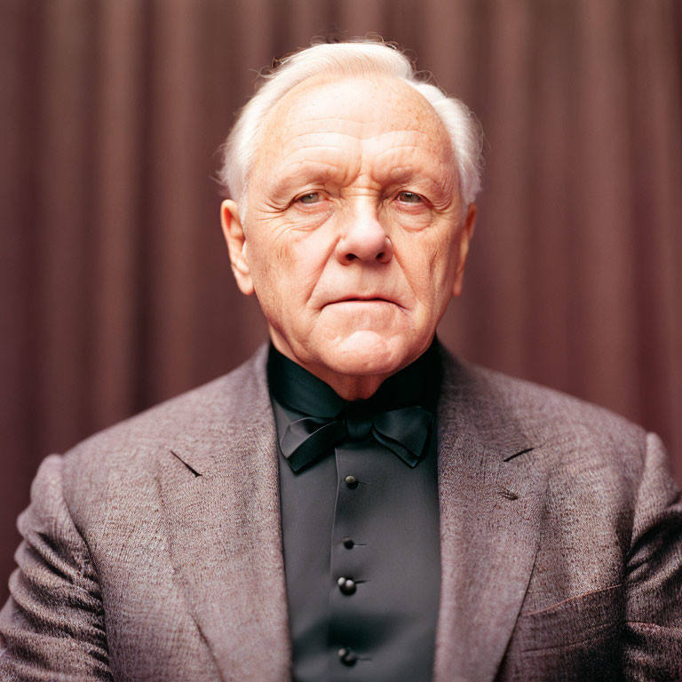 Elderly Man in Grey Suit with Black Bow Tie Against Brown Curtain Background