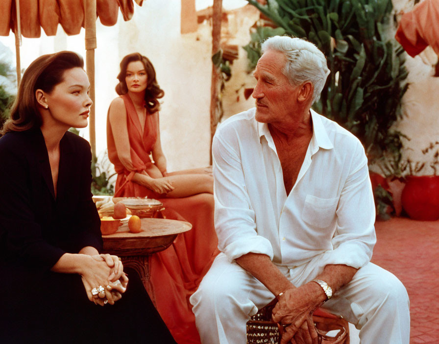 Man in white suit faces woman in black dress, observed by woman in salmon dress