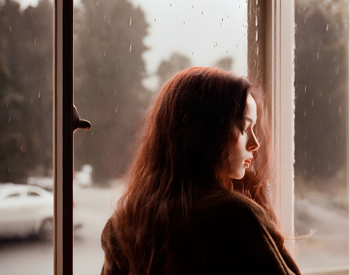 Person looking out rain-spattered window under warm light