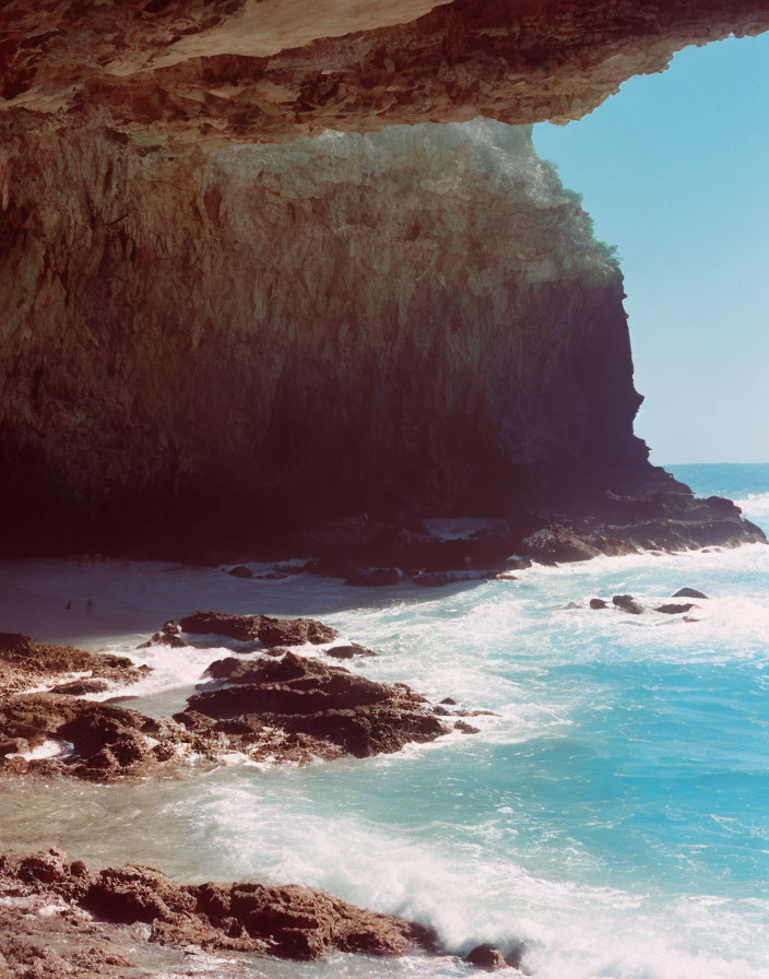 Rocky seaside overhang with sandy beach, boulders, and distant figures.