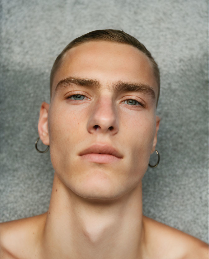 Young man with blue eyes and hoop earrings on gray background