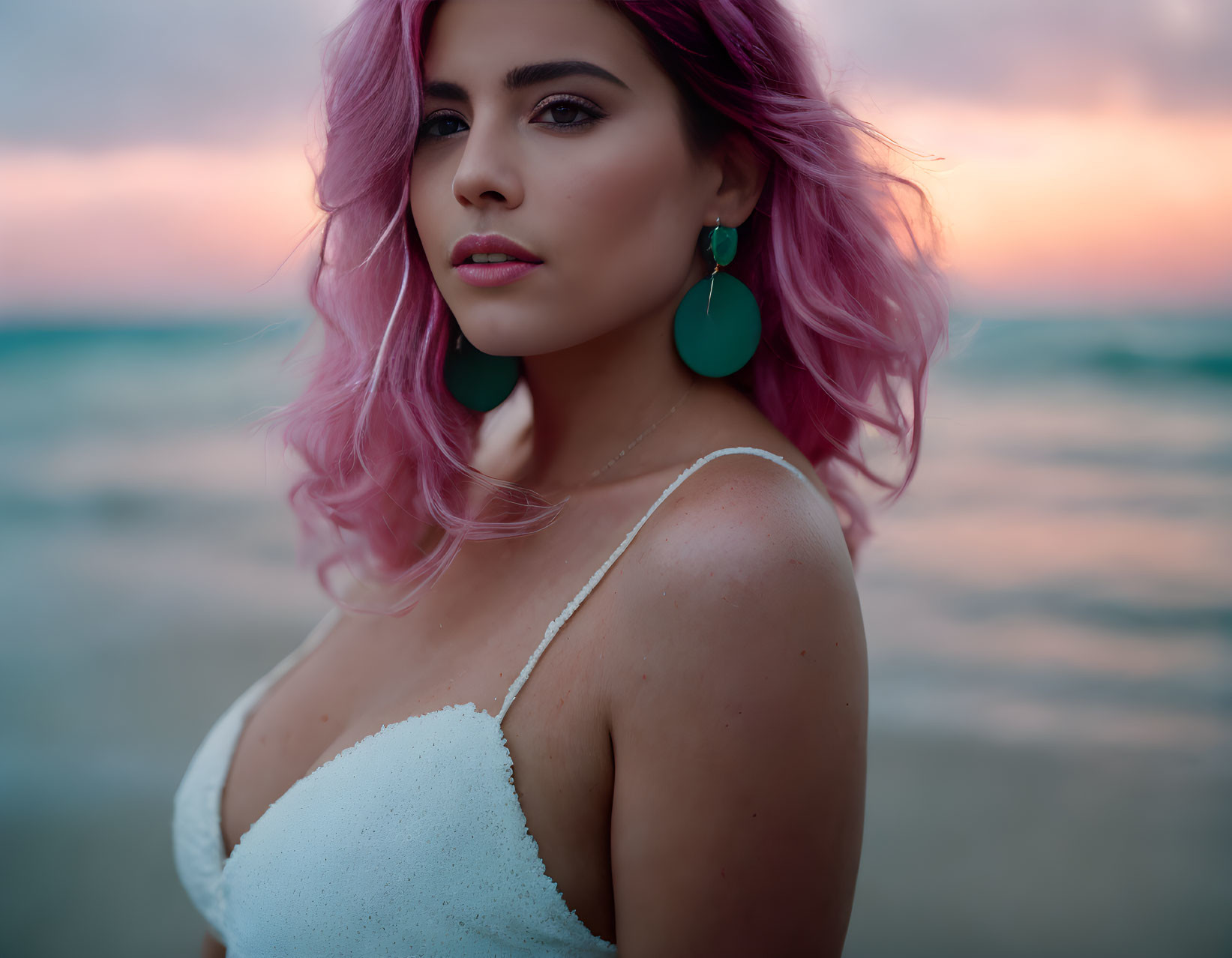 Pink-haired woman with teal earrings against beach sunset.