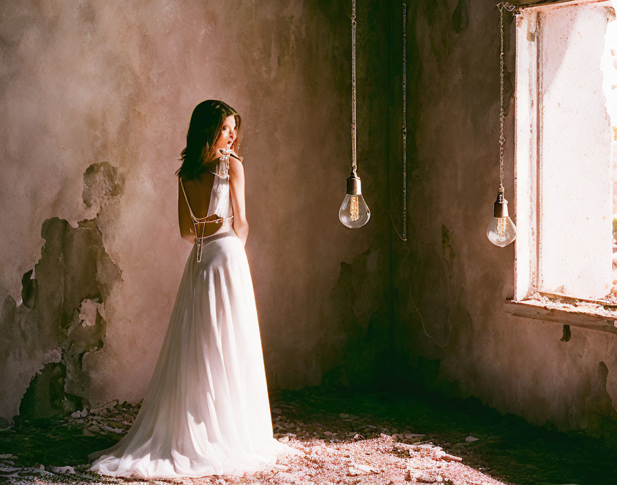 Woman in white gown in sunlit room with peeling walls and hanging light bulbs