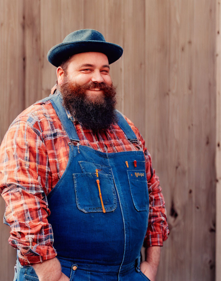 Bearded man in bowler hat and denim overalls poses against wood-panel background