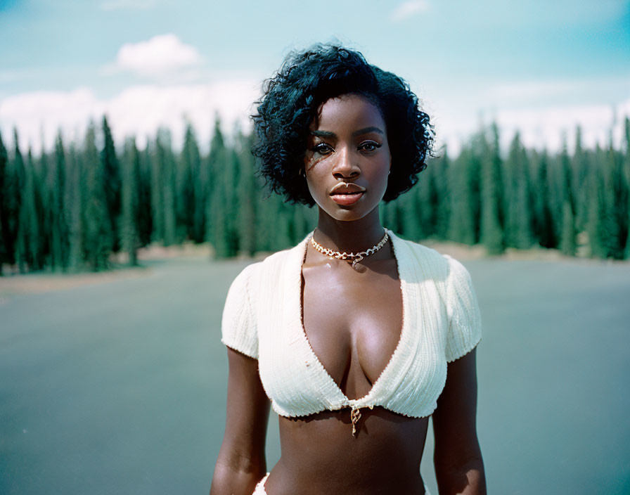 Woman with Short Curly Hair in White Crop Top and Necklace Against Pine Trees and Blue Sky