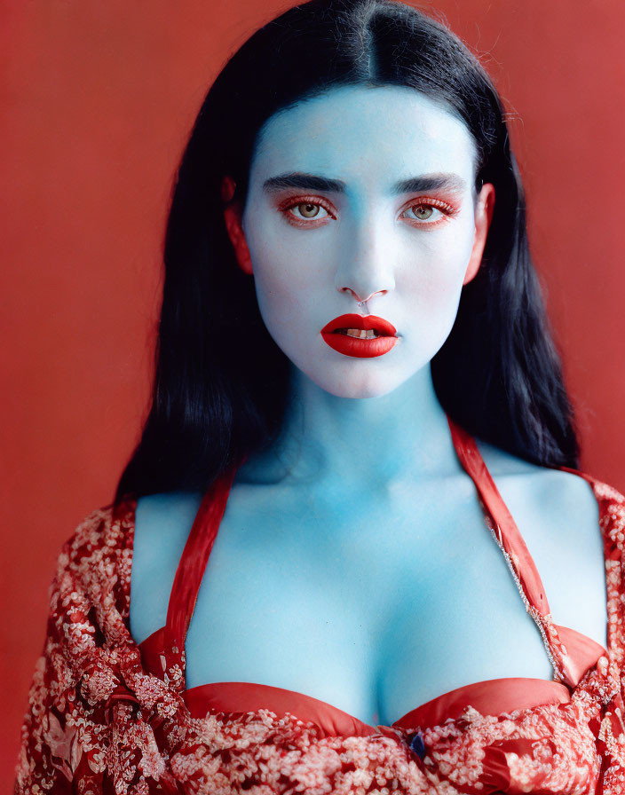 Blue-skinned woman with dark hair and red makeup in floral top on red background