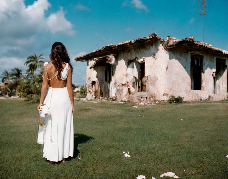 Woman in white dress holding hat near dilapidated building under clear blue sky