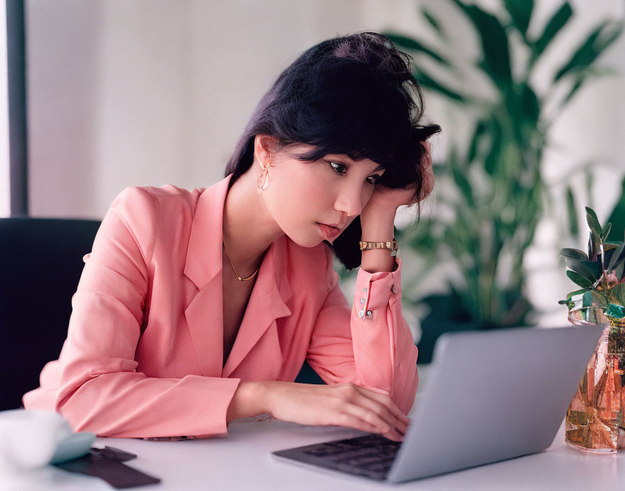 Professional woman in pink blazer working on laptop at white desk