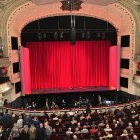 Theater interior with red curtains, green ornate seating, and audience.