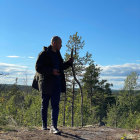 Character with backpack and weapons overlooking forested valley with spear in hand in serene video game landscape