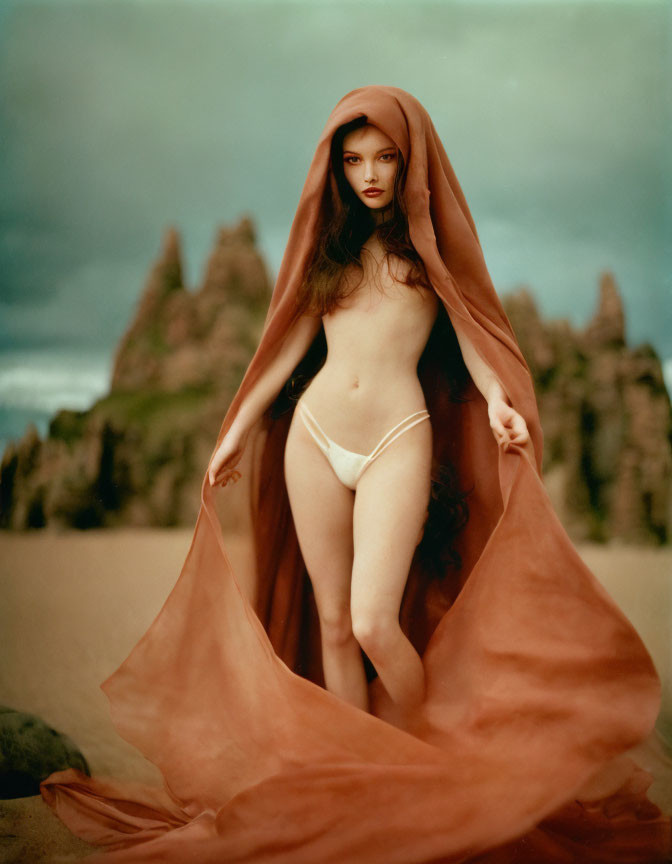 Woman in terracotta cloak on sandy terrain with jagged rocks under stormy sky