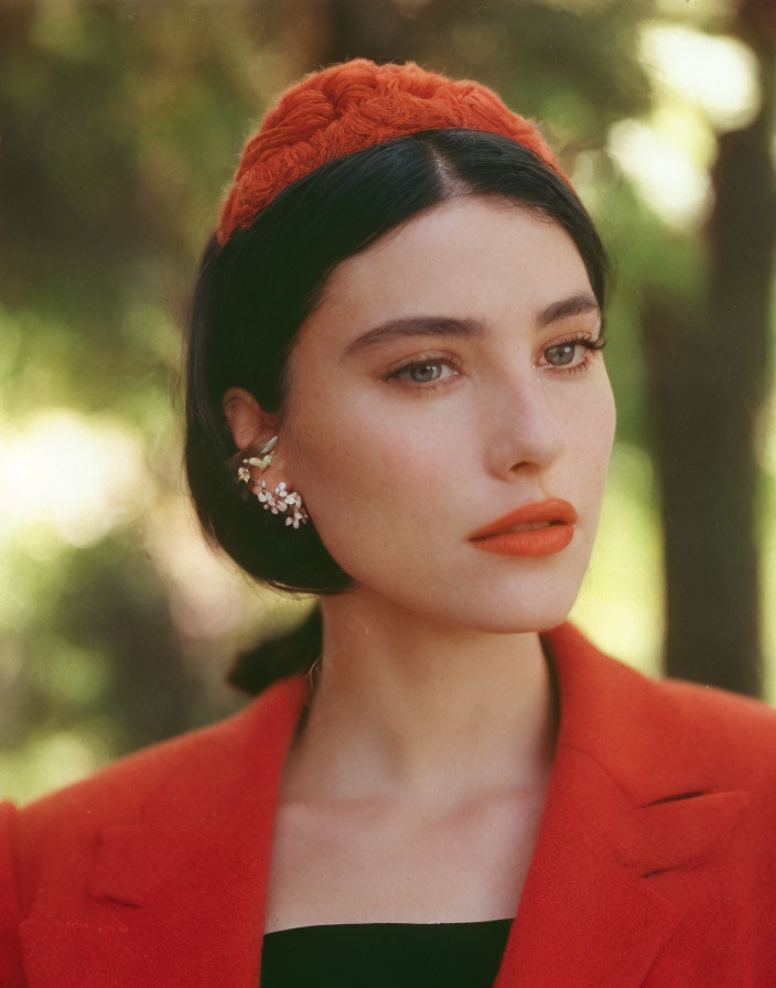 Dark-haired woman in orange hat, red blazer, and floral earring on green background