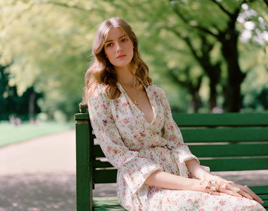 Young woman with wavy hair in floral dress sitting on park bench surrounded by green trees