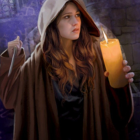 Woman in hooded cloak with lit candle and matchstick against medieval stone wall.