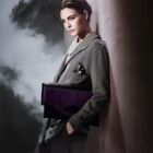 Elegant woman in gray suit with fur collar holding purple handbag poses against geometric background