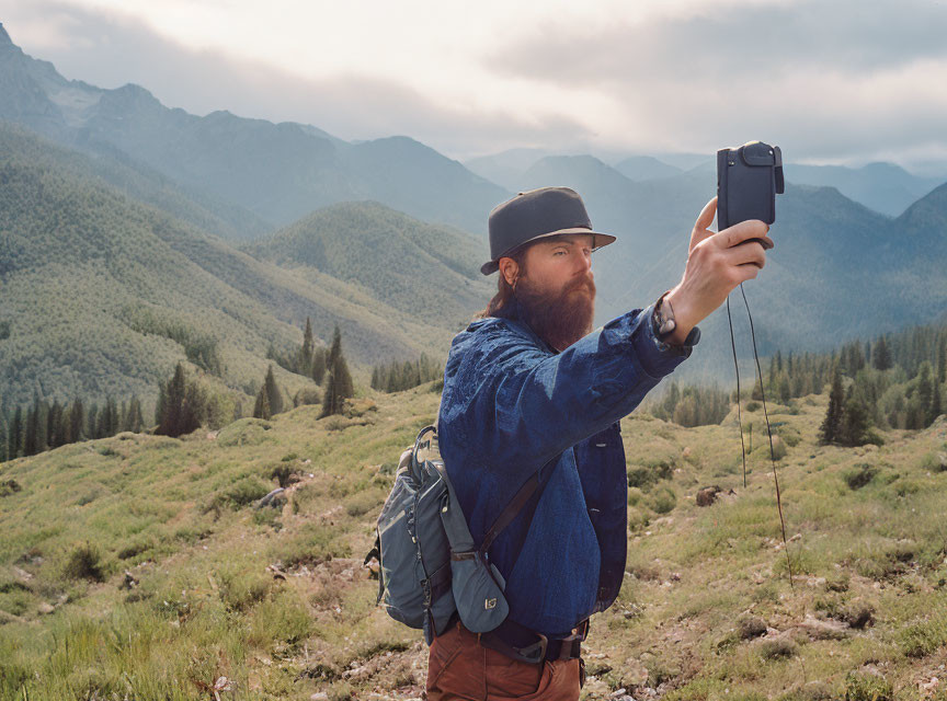 Bearded man in hat takes selfie in mountainous landscape