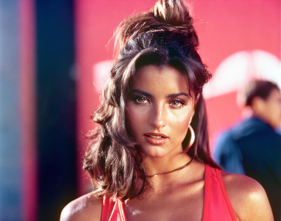 Woman with high ponytail in dramatic makeup wearing red top against blurred blue and red background