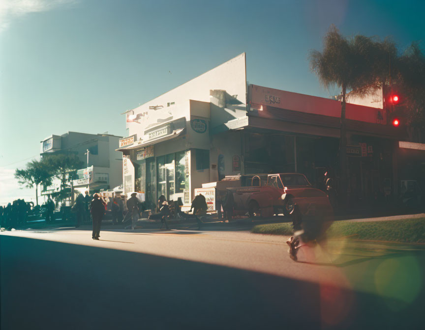 Vintage cars and pedestrians on sunlit street with storefronts and traffic lights under clear sky, featuring lens