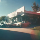 Vintage cars and pedestrians on sunlit street with storefronts and traffic lights under clear sky, featuring lens