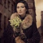 Stylish woman with voluminous curls in green sunglasses and fur-trimmed coat by snowy window