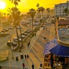 Scenic coastal road at sunset with palm trees, parked cars, beach houses, and serene sea