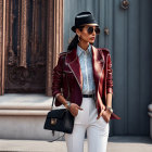 Fashionable woman in black cap and sunglasses with maroon jacket and handbag leaning against wall with curtains