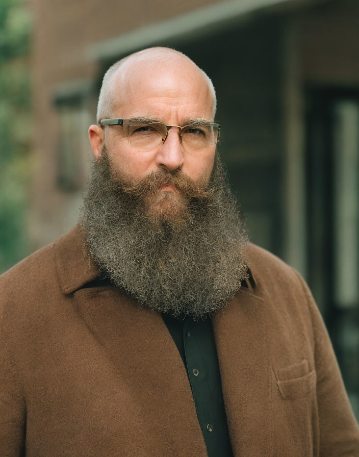 Bearded man in glasses wearing brown coat over black shirt gazes at camera