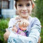 Young girl in pink beret and white cardigan holds flower-adorned doll against nature backdrop