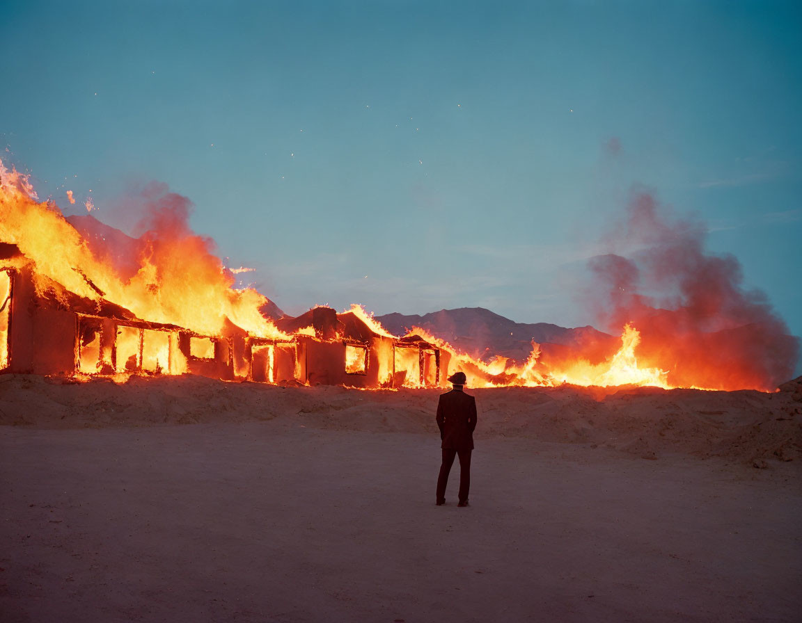 Person standing before row of buildings engulfed in flames at twilight.