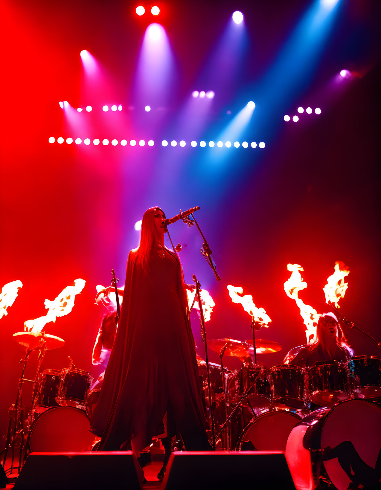 Long-haired musician performs on stage under red lights with flames and drummer visible