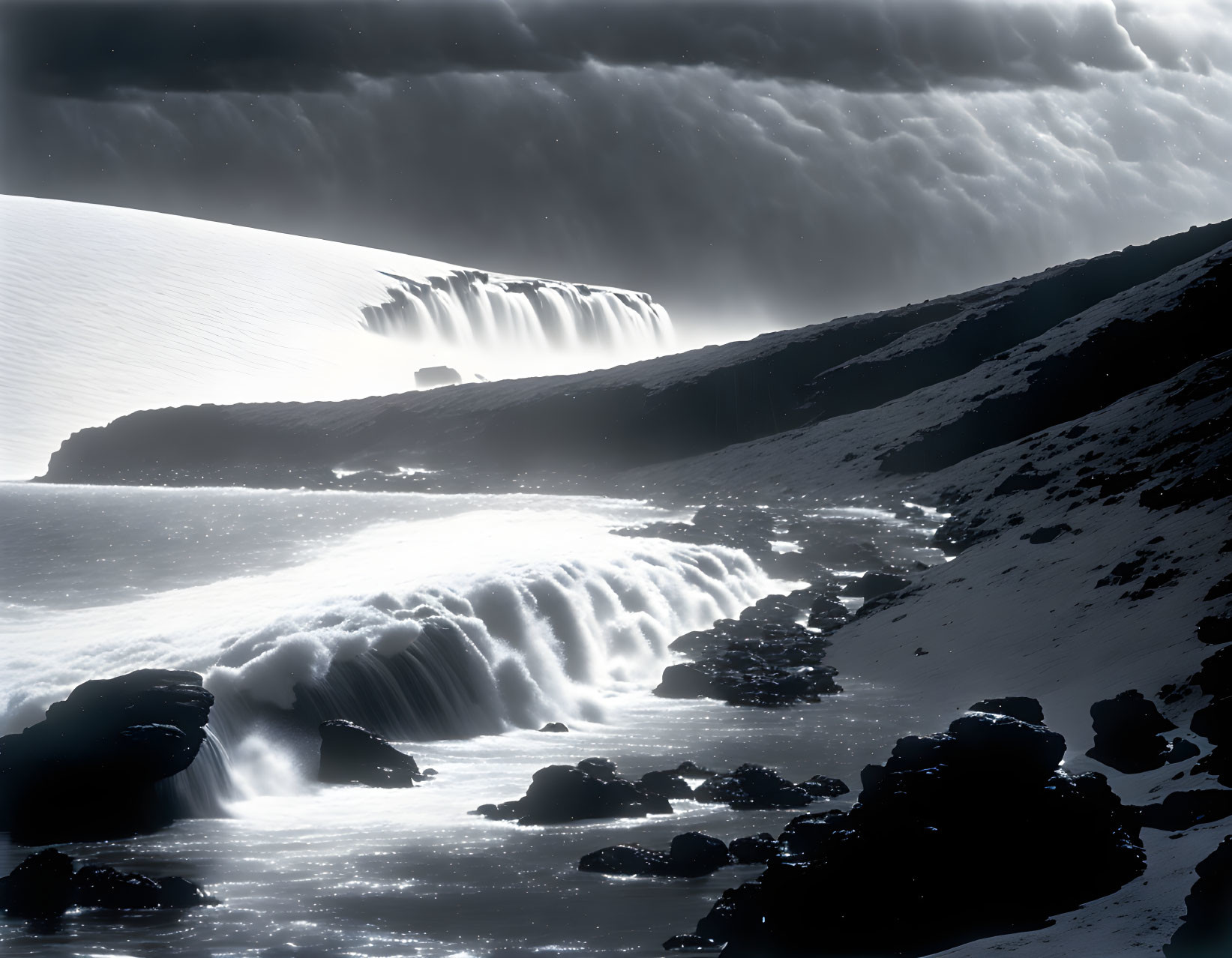 Monochrome landscape with cascading water, snow-covered terrain, rocky shoreline, dramatic clouds