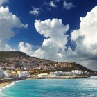 Historical ships by bustling beach with fortified city and towering clouds.