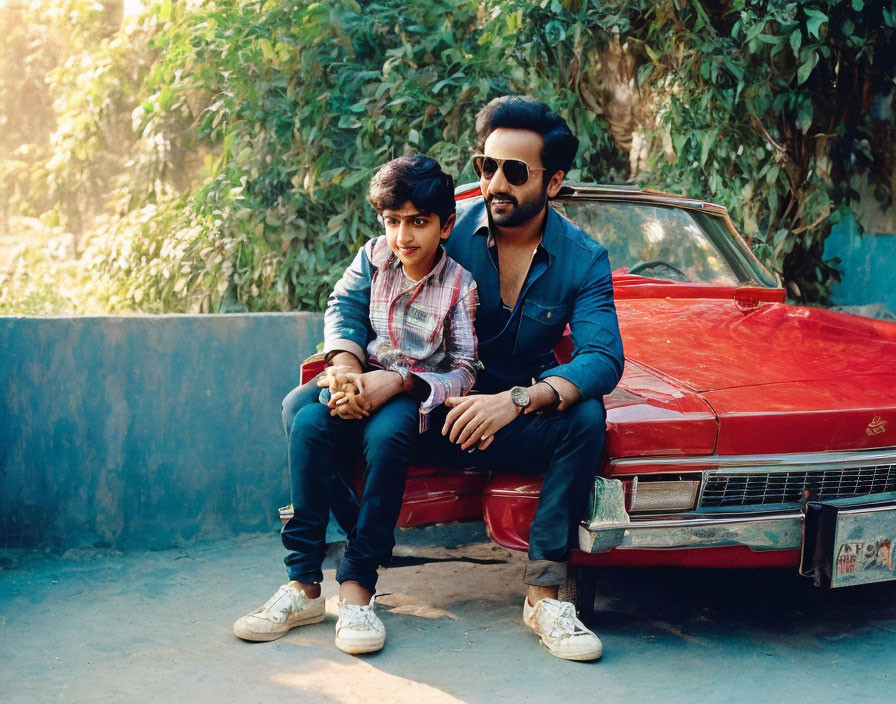 Two people on hood of red vintage car: one in checkered shirt, other in dark jacket and