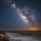 Star-studded night sky transitions to dawn over serene ocean with waves and rocky outcrops