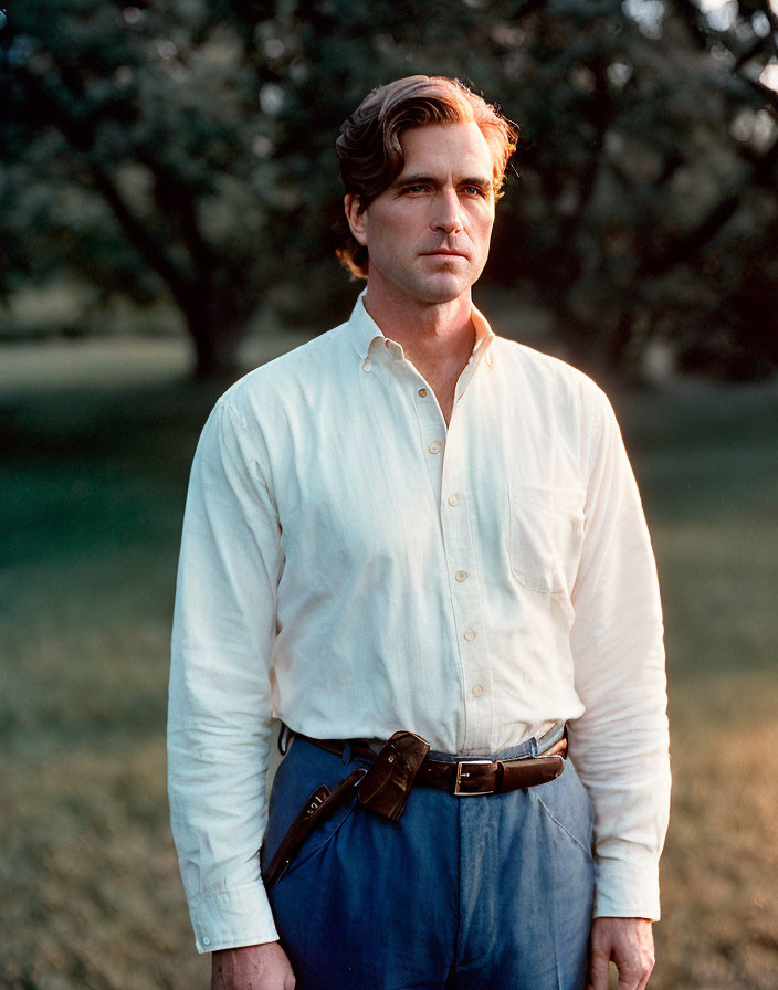 Man in white shirt and dark trousers standing in field with trees in golden hour light