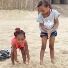 Two young girls playing on a beach, one crouching, the other standing, smiling at the