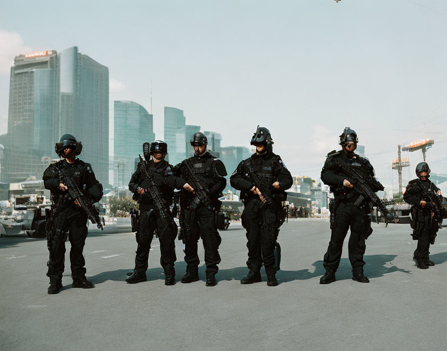 Riot police in full gear on city street with skyscrapers & cranes.