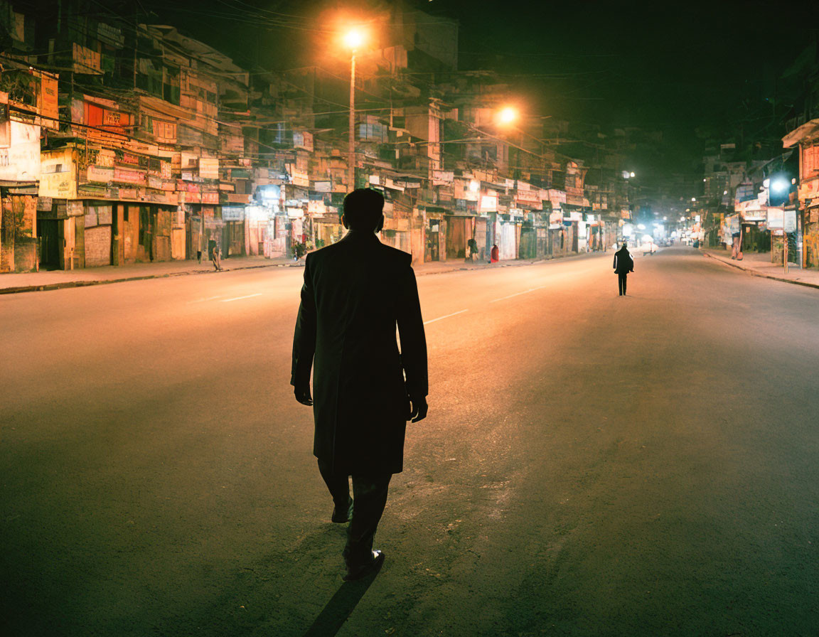 Man in coat walking down urban street at night with illuminated buildings.