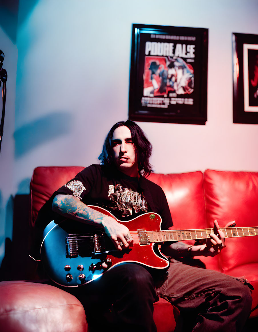 Tattooed musician playing red electric guitar on leather couch