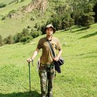 Vintage explorer in lush green field with backpack observing hill and trees under clear sky