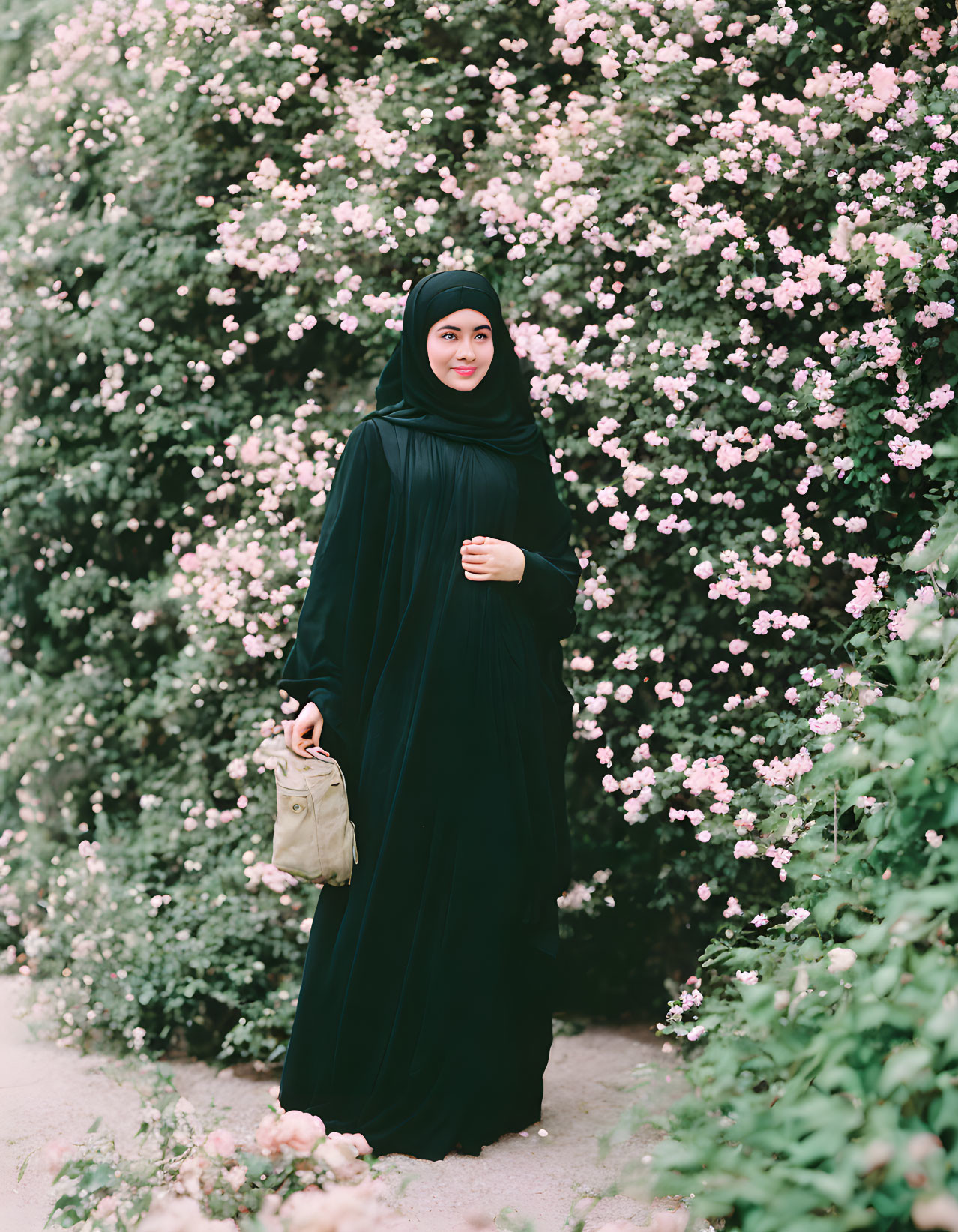 Woman in black hijab and abaya with tan bag among pink flowers