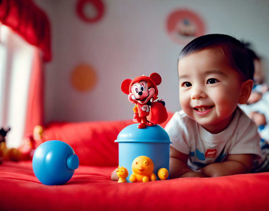 Happy baby with Mickey Mouse toy and duck-themed container on red surface