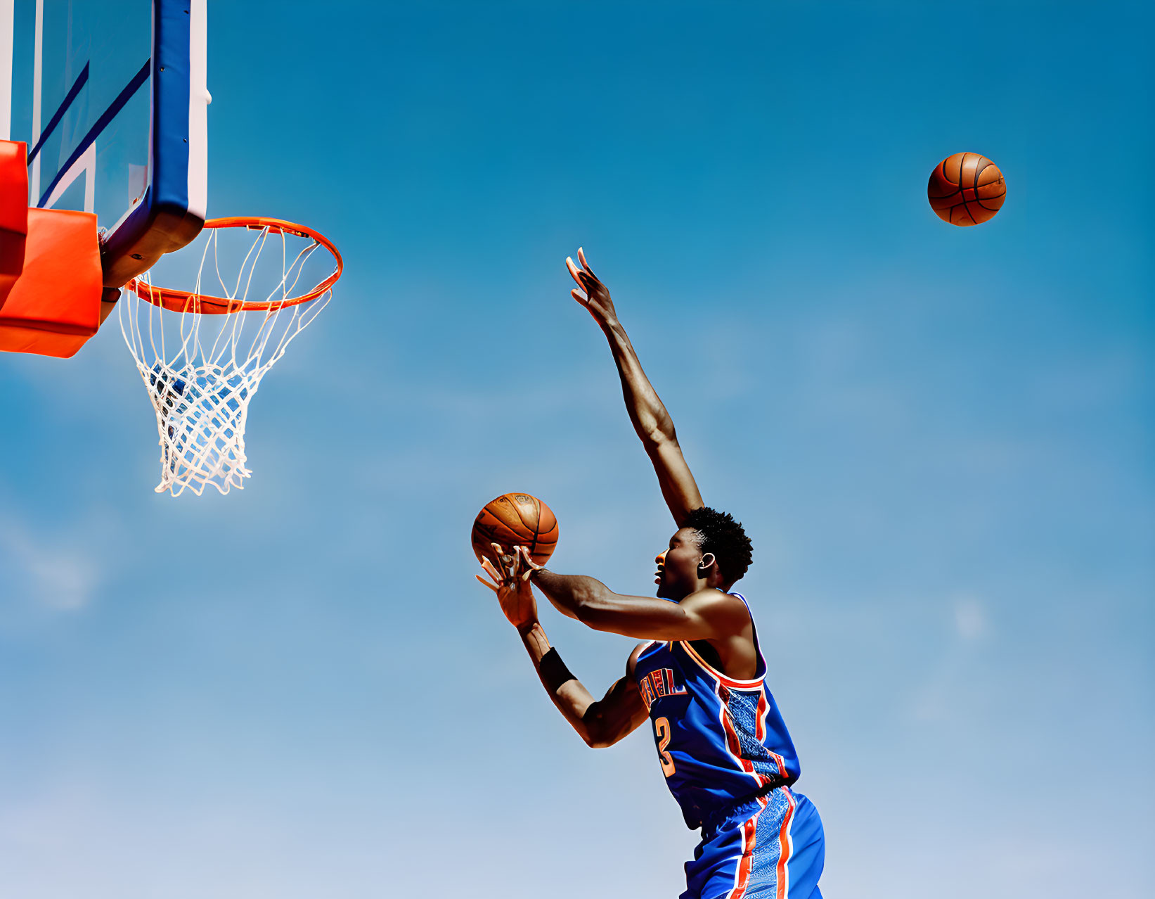 Basketball player in blue and orange attire shooting ball under clear blue sky