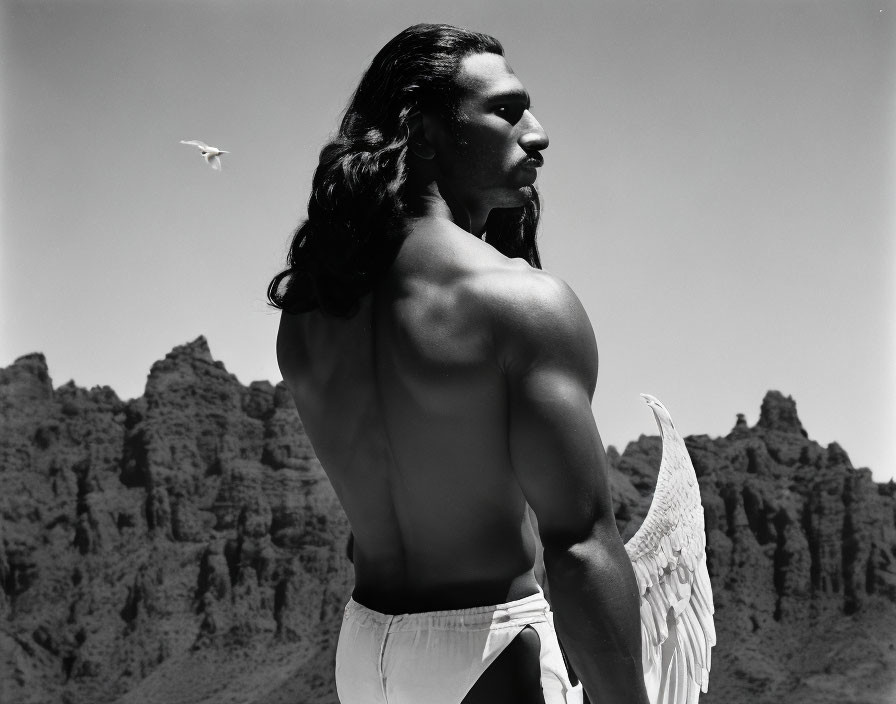 Man with feathered wings standing shirtless against rocky backdrop