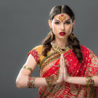 Traditional Indian woman in Namaste pose, wearing gold jewelry on grey background